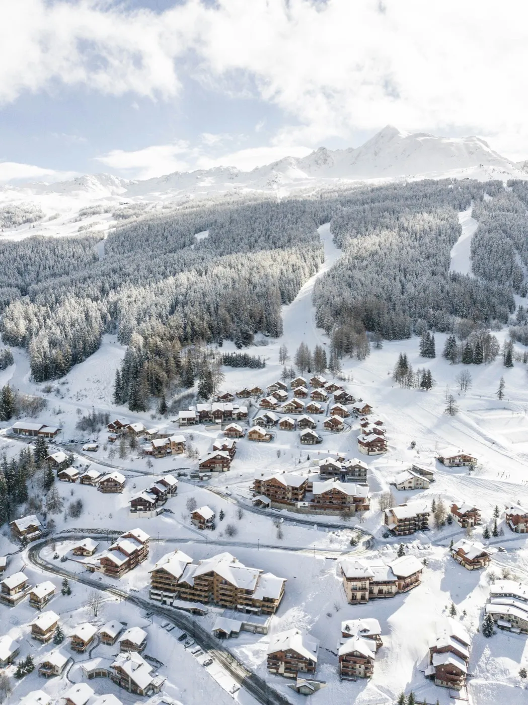 Aerial view of a snow-covered ski resort village with clustered chalets and ski runs rising into the mountains